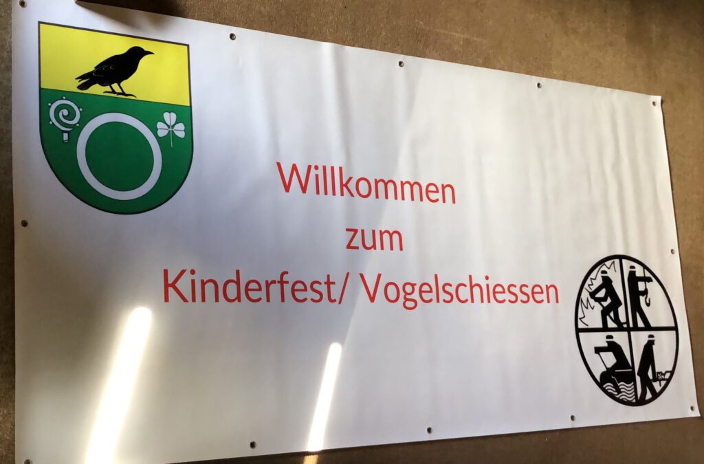 Das Bild zeigt ein weißes Plakat mit dem Wappen der Gemeinde Warnau oben links, dem allgemeinen Feuerwehrlogo unten rechts und mittig zentriert den Text "Willkommen zum Kinderfest / Vogelschiessen".